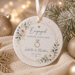 Op de afbeelding: Een rond wit keramieken ornament hangend aan een crèmekleurig lint. Het ontwerp bevat een gouden geometrisch frame, bloemaccenten en de tekst Engaged, FOREVER STARTS NOW, Isabella & Matthew, 02.14.2026 met een gouden ringillustratie.
