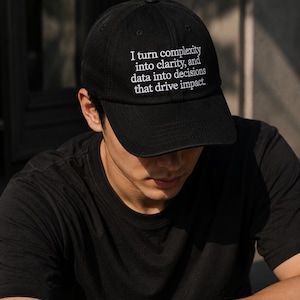 May include: Black baseball cap with white embroidered text: "I turn complexity into clarity, and data into decisions that drive impact." The cap is worn by a person wearing a black t-shirt and a silver wristwatch.
