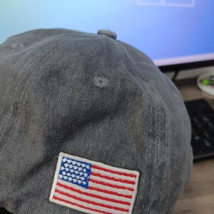 Puede incluir: Gorra de b&eacute;isbol gris con un parche bordado de la bandera estadounidense. La bandera presenta rayas rojas y blancas y un campo azul con estrellas blancas. La gorra tiene una visera curva y un bot&oacute;n en la parte superior.