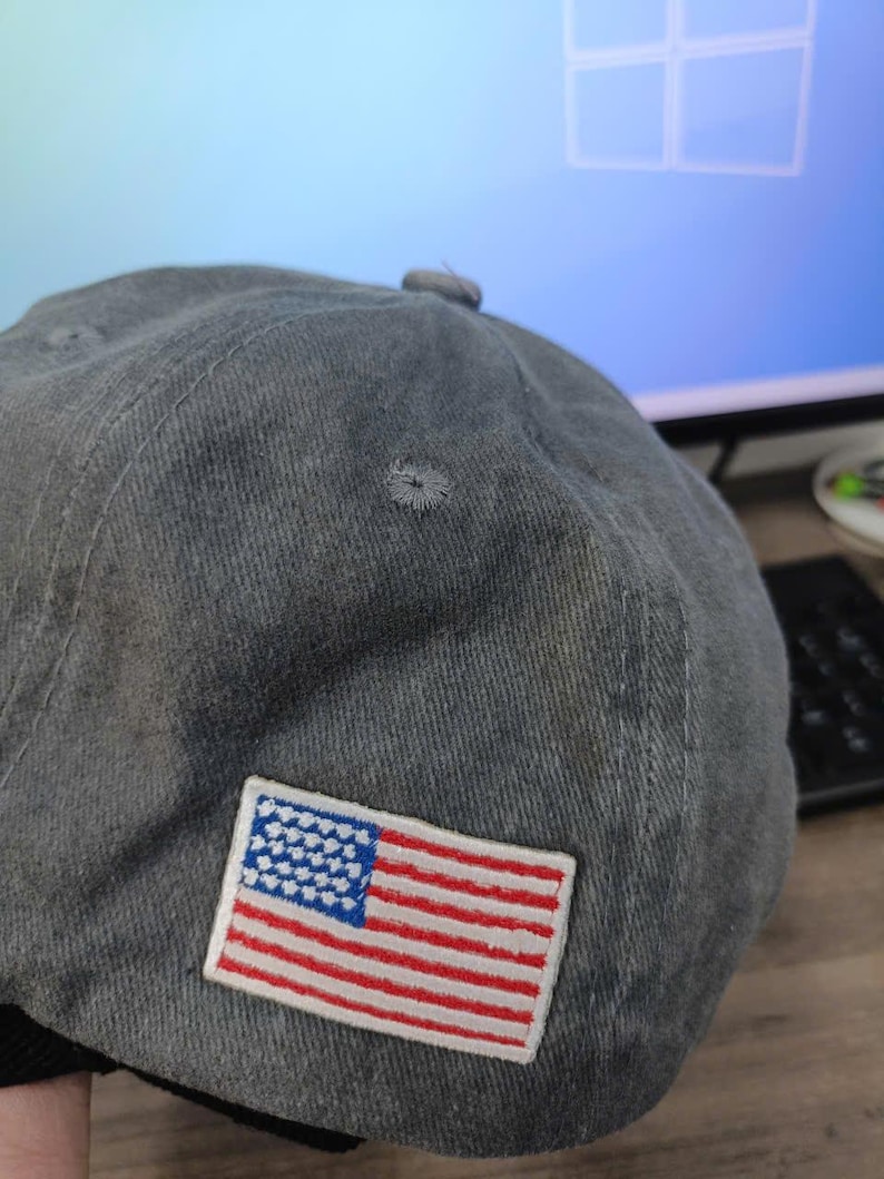 Puede incluir: Gorra de b&eacute;isbol gris con un parche bordado de la bandera estadounidense. La bandera presenta rayas rojas y blancas y un campo azul con estrellas blancas. La gorra tiene una visera curva y un bot&oacute;n en la parte superior.
