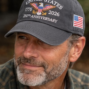 Puede incluir: Gorra de b&eacute;isbol gris oscuro con texto bordado en blanco "UNITED STATES 1776 2026 250TH ANNIVERSARY". La gorra presenta un dise&ntilde;o de &aacute;guila bordada y una peque&ntilde;a bandera americana en el lateral.