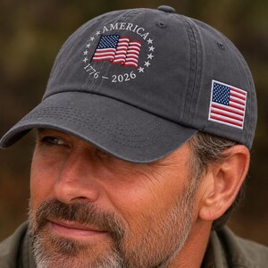 May include: A charcoal grey baseball cap featuring an embroidered design on the front with a waving American flag, the word AMERICA, and the years 1776 - 2026 encircled by stars. A small rectangular American flag patch is stitched on the side panel.