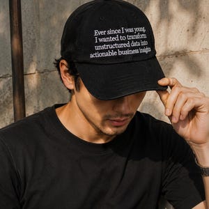 Puede incluir: Gorra de béisbol negra con texto blanco: "Desde que era joven, quería transformar datos no estructurados en información comercial procesable."