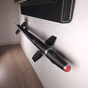 Può includere: Un oggetto cilindrico nero con piccole ali e una punta rossa, montato su un muro bianco. L'oggetto ha una fascia argentata e un'estremità appuntita. Lo sfondo include una soundbar nera e un pavimento in legno.