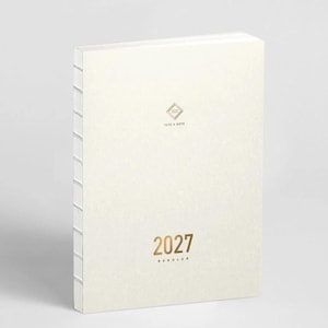 May include: A cream-colored planner with an exposed stitched spine. The cover features a small gold geometric logo above the text "TAKE A NOTE" and the year "2027" printed in gold below, followed by the word "REGULAR" in smaller gold text.