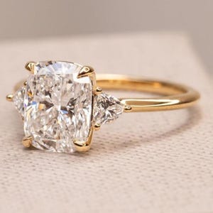 May include: A gold ring with a large, rectangular-cut diamond centre stone, and two smaller, triangular diamonds. The ring's band is polished gold. The diamonds sparkle against a neutral background.