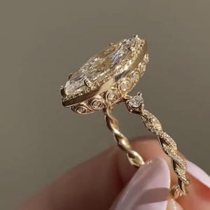 Op de afbeelding: Een gouden ring met een grote marquise-geslepen middensteen omringd door een halo van kleinere ronde stenen. De zetting bevat gedetailleerd cirkelvormig metaalwerk en de band heeft een gedraaid ontwerp ingelegd met kleine pavé-diamantjes.