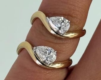 May include: Two gold rings with a pear-shaped diamond set in a swirling design. The rings are displayed on a finger against a light background. The diamonds sparkle, reflecting light.
