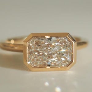 May include: A gold ring featuring a rectangular, light-coloured gemstone. The gemstone is set within a gold bezel, and the band is also gold. The ring is displayed on a white surface, with soft lighting.