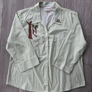 May include: A light green button-down shirt with vertical stripes. The shirt features embroidered cat designs on the front and a tree design. The shirt has long sleeves and a collared neckline. The brand name "Casolas" is visible on the label.