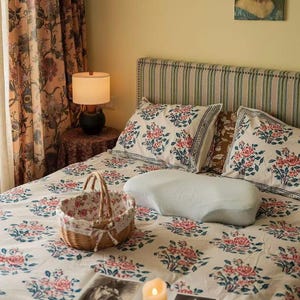 May include: A bedroom scene featuring a bed with floral patterned bedding in pink and blue. A white ergonomic pillow rests on the bed. A wicker basket with a floral lining sits on the bed, and a lit lamp is on a side table.