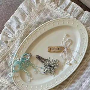 May include: Three decorative hair clips on an oval tray. One clip is gold with a beaded bow, one is silver with a starburst of clear crystals, and one is a rectangular gold clip with a dangling light blue and white beaded fringe attachment.