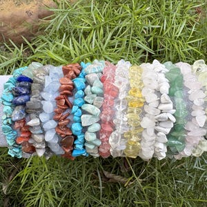 May include: A collection of gemstone chip bracelets in various colors, including turquoise, blue, red, and yellow. The bracelets are displayed on a gray cylindrical surface against a backdrop of green foliage. Each bracelet is made of small, irregular-shaped stones.