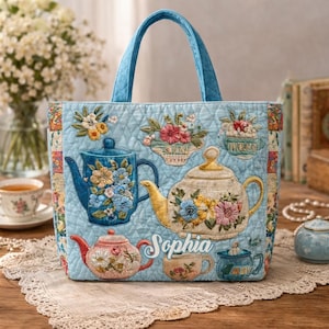 Puede incluir: Bolso tote acolchado azul claro con bordados de teteras y diseños florales. El bolso tiene un asa azul a juego y el nombre "Sophia" bordado en la parte delantera. El bolso está decorado con varias tazas de té y teteras.
