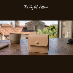 Può includere: Un porta carte in pelle beige con chiusura a bottone in ottone. Il testo "LEATHER CARD HOLDER" e "DIY Digital Pattern" sono nella parte superiore dell'immagine. Il porta carte è su un tavolo di legno.