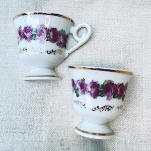 May include: Two white porcelain cups with a pedestal base, featuring a band of purple roses and green leaves around the upper body. Each cup is accented with a gold rim and delicate gold filigree patterns below the floral design. One cup has a handle.