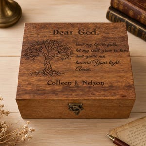 May include: A wooden box with a hinged lid and a metal clasp. The box is engraved with a tree design and the text "Dear God, root my life in faith, let my soul grow in love, and guide me toward Your light. Amen. Colleen J. Nelson."