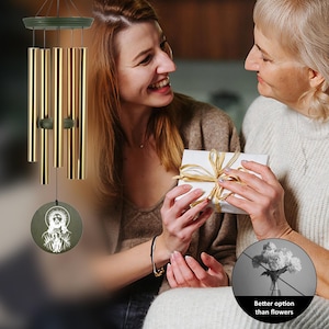 May include: A golden wind chime with a circular, engraved pendant featuring a religious figure. The chime has multiple cylindrical tubes and is suspended from a dark green top. The image includes a gift exchange with the text "Better option than flowers."