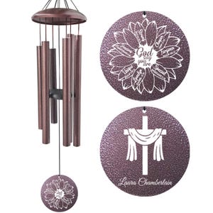 May include: A bronze-colored wind chime with a floral design and the text "God say you are." The wind chime has multiple tubes and a round pendant with a cross design. The text "Laura Chamberlain" is also visible.
