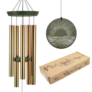 May include: A wind chime with gold-colored tubes and a dark green top and clapper. A round, dark green disc with a sun and wave design hangs nearby, with the text "HIS MERCIES ARE NEW EVERY MORNING". A cardboard box is also in the image.