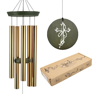 May include: A wind chime with gold-colored tubes and a dark green top and clapper. A round, dark green disc with a white cross design hangs nearby. A brown box with the brand name "Artisan" is also in the image.