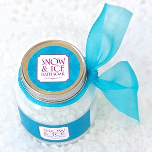 May include: A clear glass jar filled with white bath soak crystals, labeled "Snow & Ice Bath Soak." The jar has a silver lid and is adorned with a turquoise ribbon tied in a bow. The background is a white, textured surface.