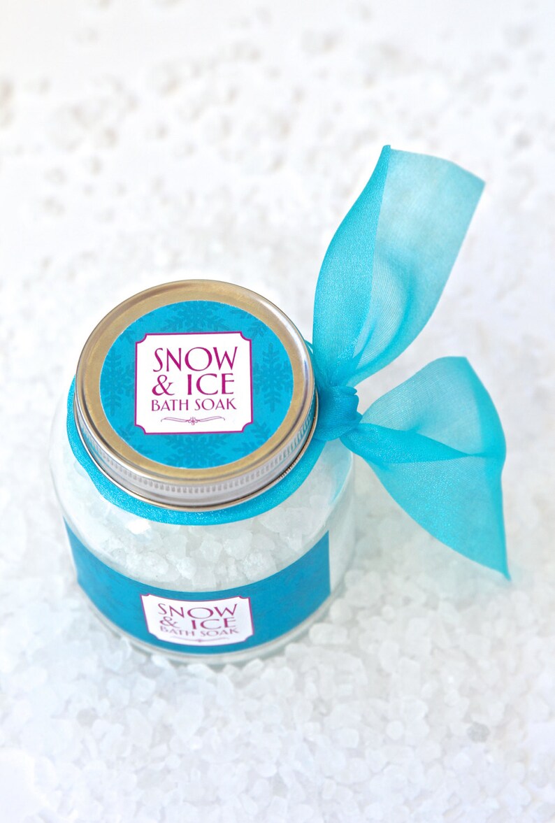 May include: A clear glass jar filled with white bath soak crystals, labeled "Snow & Ice Bath Soak." The jar has a silver lid and is adorned with a turquoise ribbon tied in a bow. The background is a white, textured surface.
