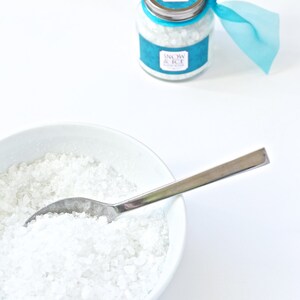 May include: A white ceramic bowl filled with coarse, white salt and a silver spoon. A glass jar with a silver lid and a teal label reading "Snow & Ice Bath Soak" is tied with a teal ribbon. The jar is also filled with white salt.
