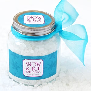 May include: A clear glass jar filled with white bath soak crystals. The jar has a silver lid and is adorned with a light blue label featuring snowflakes and the text "SNOW & ICE BATH SOAK". A matching blue ribbon is tied around the jar.