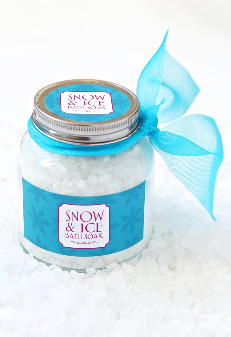 May include: A clear glass jar filled with white bath soak crystals. The jar has a silver lid and is adorned with a light blue label featuring snowflakes and the text "SNOW & ICE BATH SOAK". A matching blue ribbon is tied around the jar.