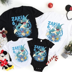 Puede incluir: Camisetas negras y blancas y un body negro con un diseño de dibujos animados azul. El diseño muestra el nombre "ZAKIA" y el número "6", junto con una criatura marina azul y tres tortugas. También se ven decoraciones festivas.