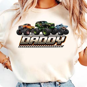 May include: Cream-colored t-shirt featuring three monster trucks in orange, green, and blue. The word "DADDY" is printed in silver with a checkered flag design underneath. The trucks have large tires.