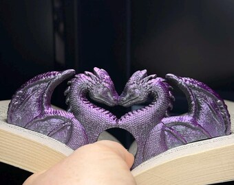 Può includere: Un fermalibri a forma di drago viola con due draghi che si fronteggiano, formando un cuore. Il fermalibri è appoggiato sulle pagine di un libro. I draghi hanno ali dettagliate e squame strutturate.