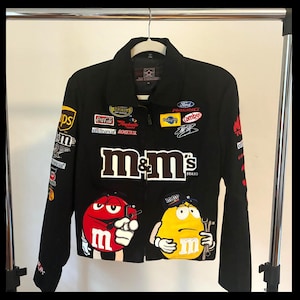 May include: Black racing jacket with multiple sponsor patches, including M&M's, Coca-Cola, and Ford Racing. The jacket features a collar, zipper closure, and cartoon M&M's characters on the front. The jacket is hanging on a metal rack.