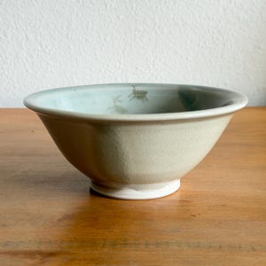 May include: A ceramic bowl with a light sage green glaze featuring a subtle crackle finish. The interior of the bowl displays a faint, hand-painted illustration of a small creature, and the base is finished with a clean, white circular foot.