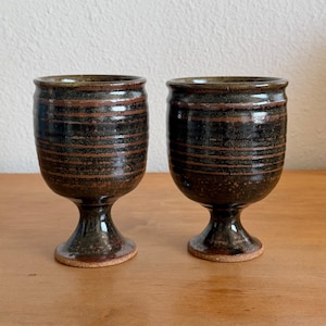 May include: Two matching ceramic goblets featuring a dark, mottled brown and black glaze with horizontal ridged bands. Each vessel has a rounded bowl, a short pedestal stem, and a circular base, showcasing a glossy finish throughout.