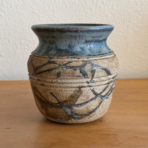 May include: A ceramic vase with a wide, flared rim featuring a smooth, glossy blue glaze. The body of the vase has a textured, sandy beige finish with dark, branching crackle patterns and horizontal incised lines encircling the midsection.