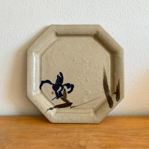 May include: An octagonal ceramic plate with a speckled beige finish. The center features a hand-painted dark blue floral motif with brown stem and leaf accents. The plate has a raised rim and a textured, matte surface throughout the design.