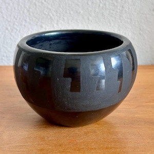 May include: A black ceramic bowl with a glossy finish. The bowl has a rounded shape with a flat base. The exterior features a matte black band with geometric shapes. The bowl is sitting on a wooden surface.