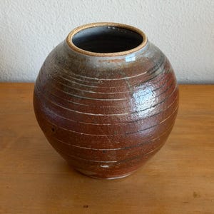 May include: A handcrafted, round ceramic vase with a wide opening. The vase features horizontal, textured lines in shades of brown and reddish-brown, with a lighter rim. The interior is dark, and the vase sits on a wooden surface.