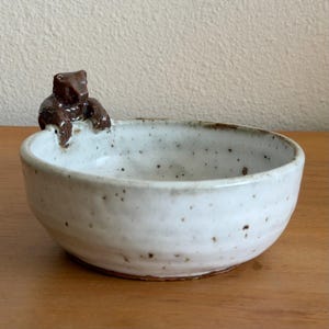 May include: A round ceramic bowl with a speckled off-white glaze and a dark brown sculpted bear figure perched on the rim. The bowl features a smooth interior and a textured exterior surface with visible brown clay flecks throughout the glaze.