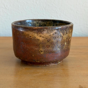 May include: A small, round ceramic bowl featuring a mottled glaze with deep reddish-brown and earthy golden-tan tones. The surface has a textured, rustic appearance with subtle variations in color and a smooth, glossy finish along the rim.