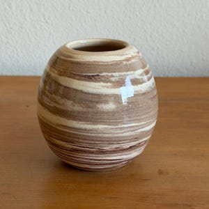 May include: A small, round ceramic vase with a wide opening. The vase features horizontal bands of brown, tan, and cream colors, creating a striped pattern. The vase is sitting on a wooden surface.