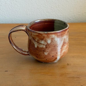 May include: A ceramic mug with a rounded body and a curved handle. The exterior features a mottled glaze with earthy tones of reddish-brown, cream, and tan, showing a speckled texture. The interior is glazed in a deep, glossy reddish-brown hue.