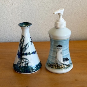 May include: Two ceramic decorative bottles. One is a white vase with blue floral and leaf designs. The other is a soap dispenser with a white pump and a lighthouse design with blue and white stripes.