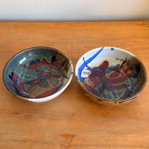 May include: Two handmade ceramic bowls with a unique design. One bowl features a dark blue and brown background with red leaf-like shapes. The other bowl has a white and tan background with red and blue abstract designs. Both bowls have a glossy finish.