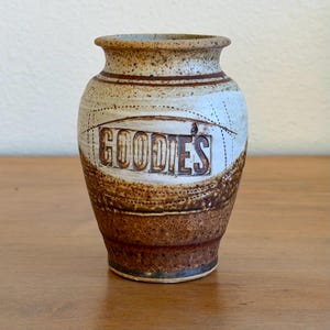 May include: A ceramic vase with a speckled beige and brown glaze. The word GOODIES is embossed in a rectangular frame on the front. The vase features a flared rim, a rounded body, and a tapered base with horizontal incised line details.