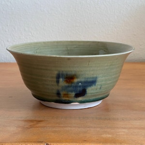 May include: A ceramic bowl with a light sage green glaze featuring a fine crackle finish. The exterior displays a prominent horizontal band of dark blue and burnt orange abstract markings. The bowl has a wide, flared rim and a white base.