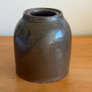 May include: A brown stoneware jar with a lid. The jar has a rounded body and a slightly flared rim. The exterior has a glossy finish with variations in color. The jar is sitting on a wooden surface.
