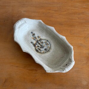 May include: An oblong ceramic tray with a speckled off-white glaze and scalloped edges. The center features a dark gray and amber floral-inspired motif. The tray includes two small, ribbed handles on opposite ends, creating a textured finish.